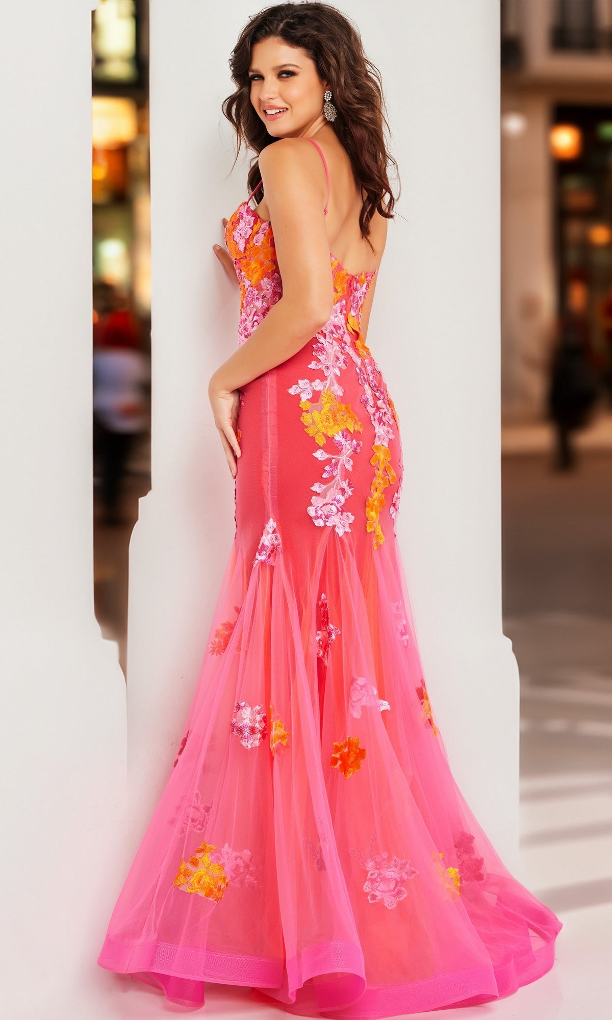 A woman wearing the Long Prom Dress 36843 by Jovani—a bright pink mermaid tulle gown with yellow and white floral embroidery—stands against a white pillar, looking back and smiling.